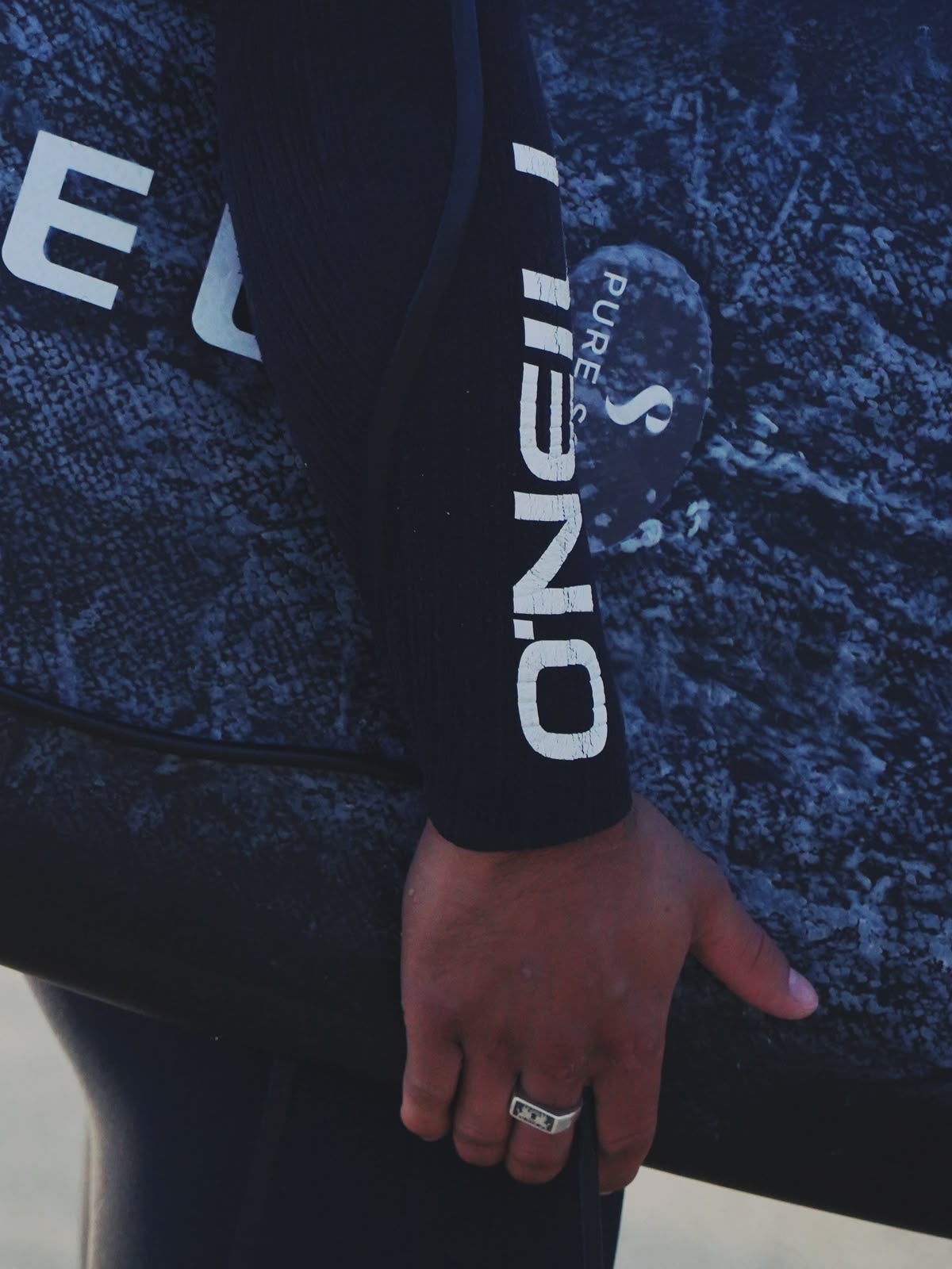 Surfer in a wetsuit carrying a board branded with the Pure Scot mark