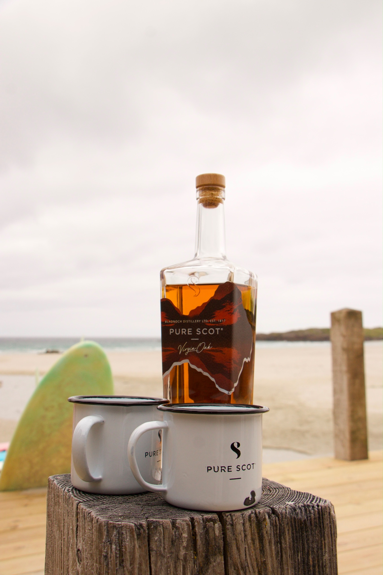 Pure Scot whisky bottle and two branded enamel mugs on a driftwood post beside the beach