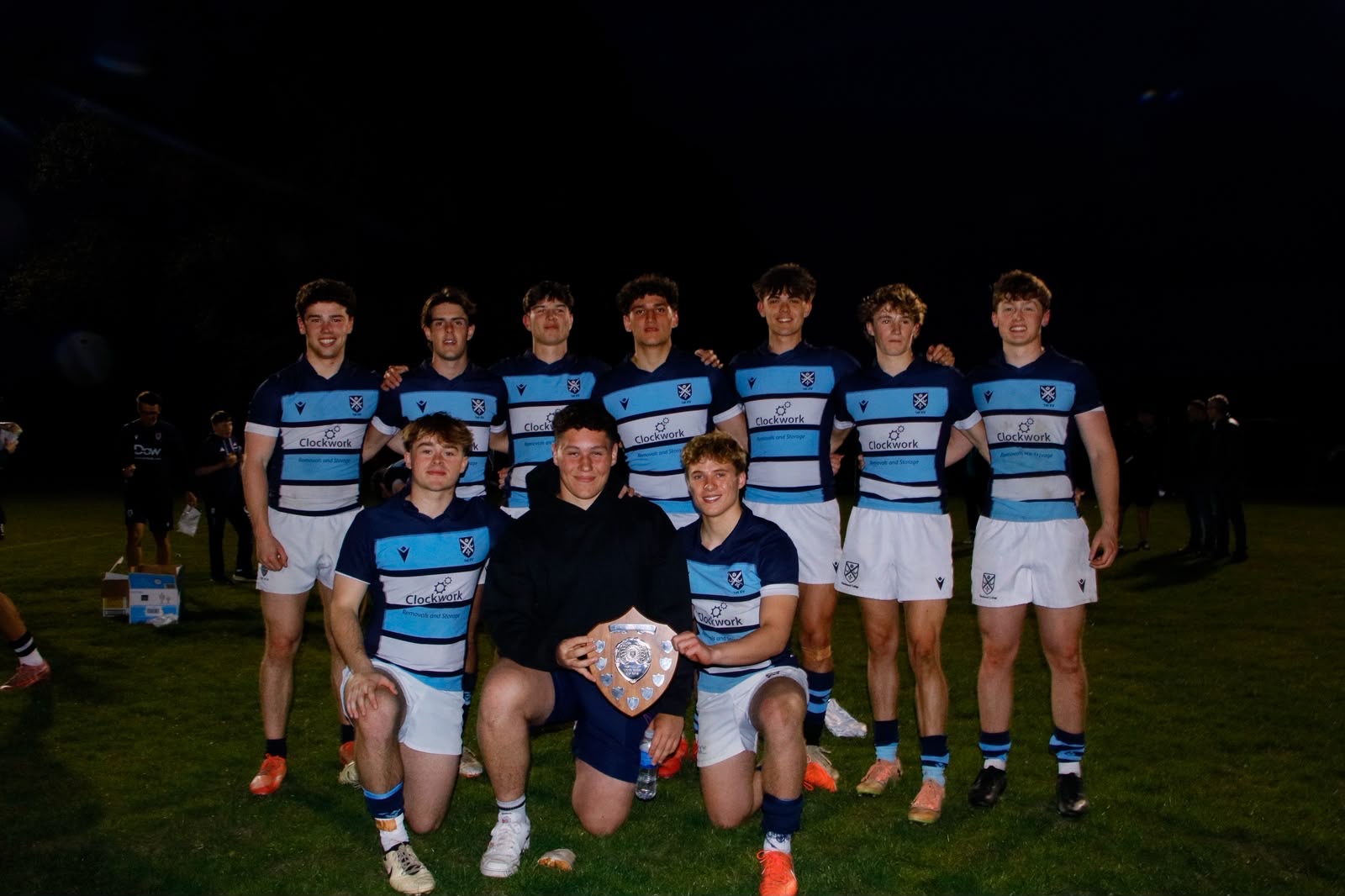 Rugby squad in blue and white kit with their trophy on the pitch at night