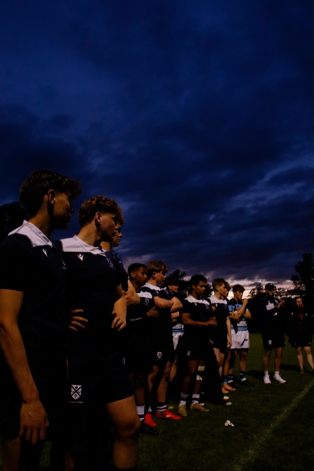 Rugby players lined up on the pitch at dusk under a dark sky