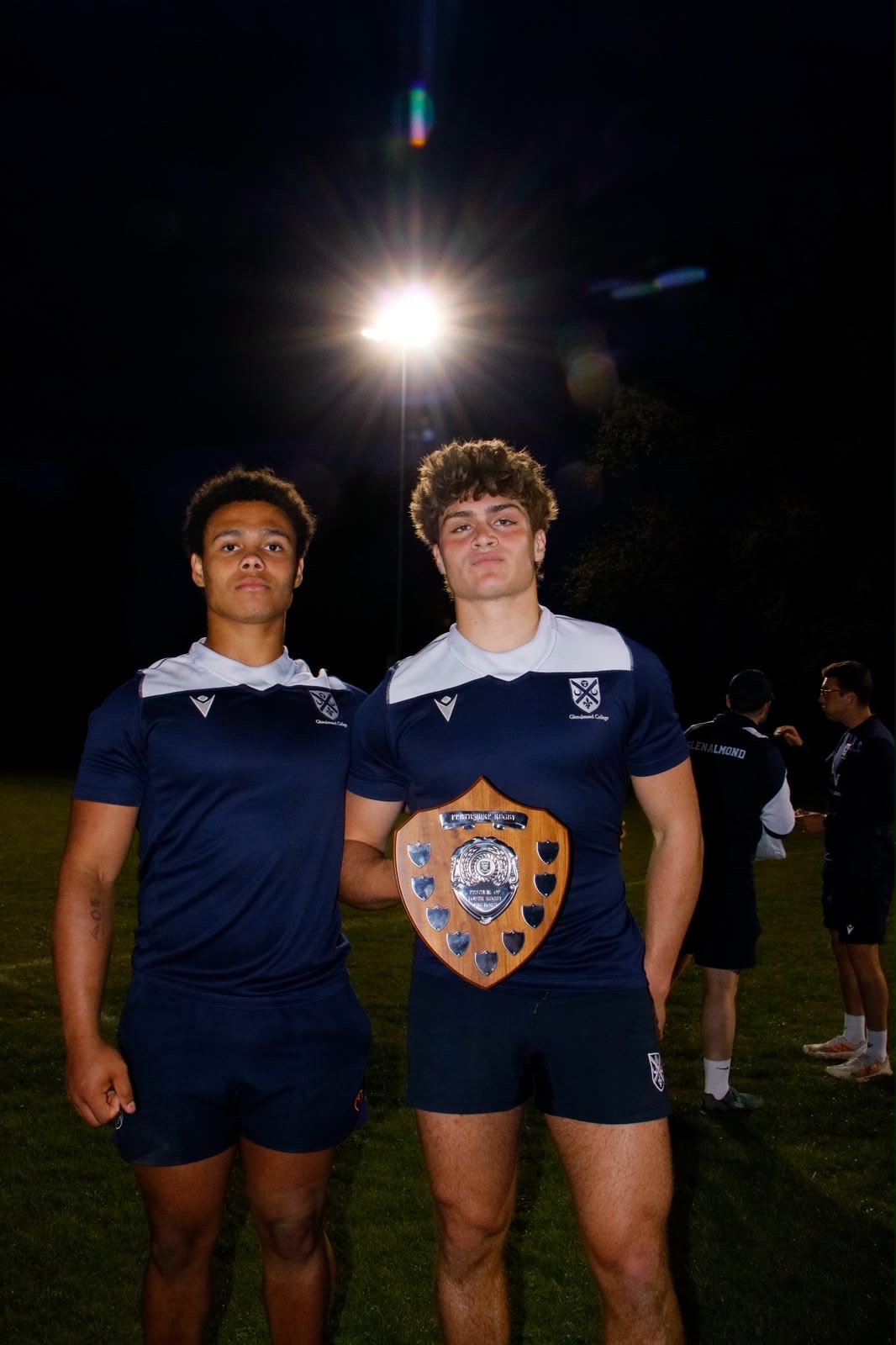 Two rugby players holding a wooden trophy shield at night under stadium lights