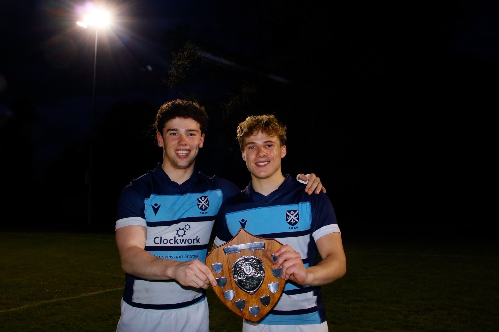 Two players in blue and white jerseys holding the winners' shield under floodlights